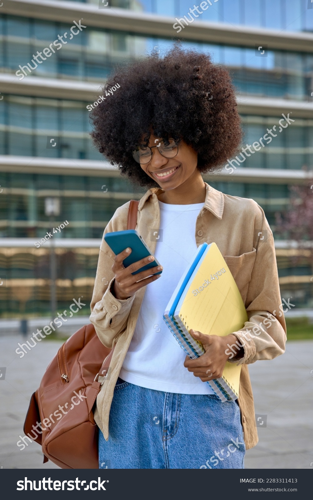 Vertical portrait of happy African American student girl enjoy online communication, hold exercise books with a backpack, scroll social media app on cell phone device on university campus background. #2283311413