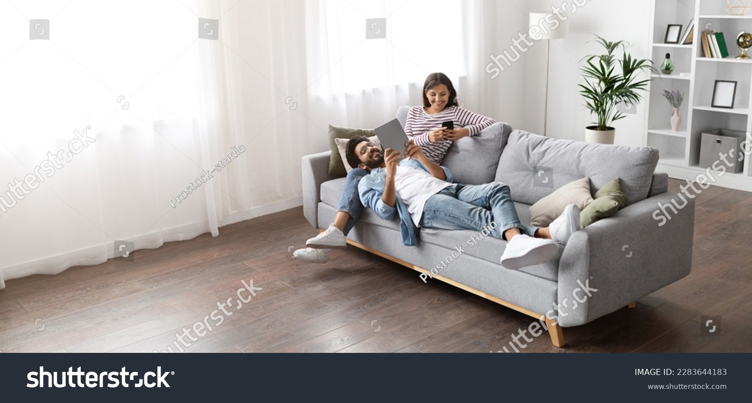 High angle view of happy smiling multiracial millennial couple relaxing together on couch at home, handsome arab guy wearing glasses using digital tablet lying on indian lady laps, copy space, banner #2283644183