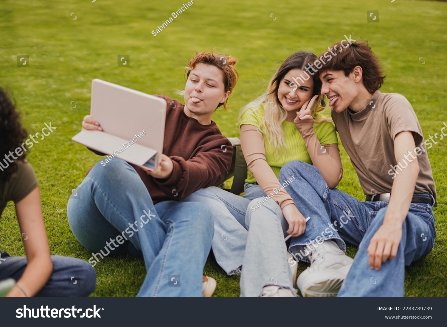 Young students having fun together taking a selfie with digital tablet while sitting outside of university meadow #2283789739