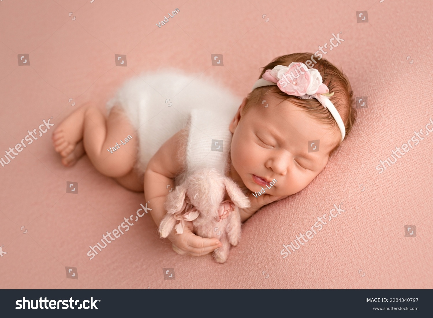 Top view of a newborn baby girl sleeping in a white jumpsuit with pink rabbit a white bandage and a pink flower on her head on a pink background. Beautiful portrait of a little girl 7 days, one week. #2284340797