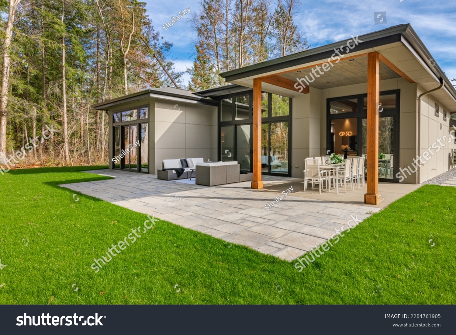 Exterior image of a contemporary home with flat roof and brown trim and lush green grass blue sky forest in the background furnished patio for outdoor dining and lounging #2284761905