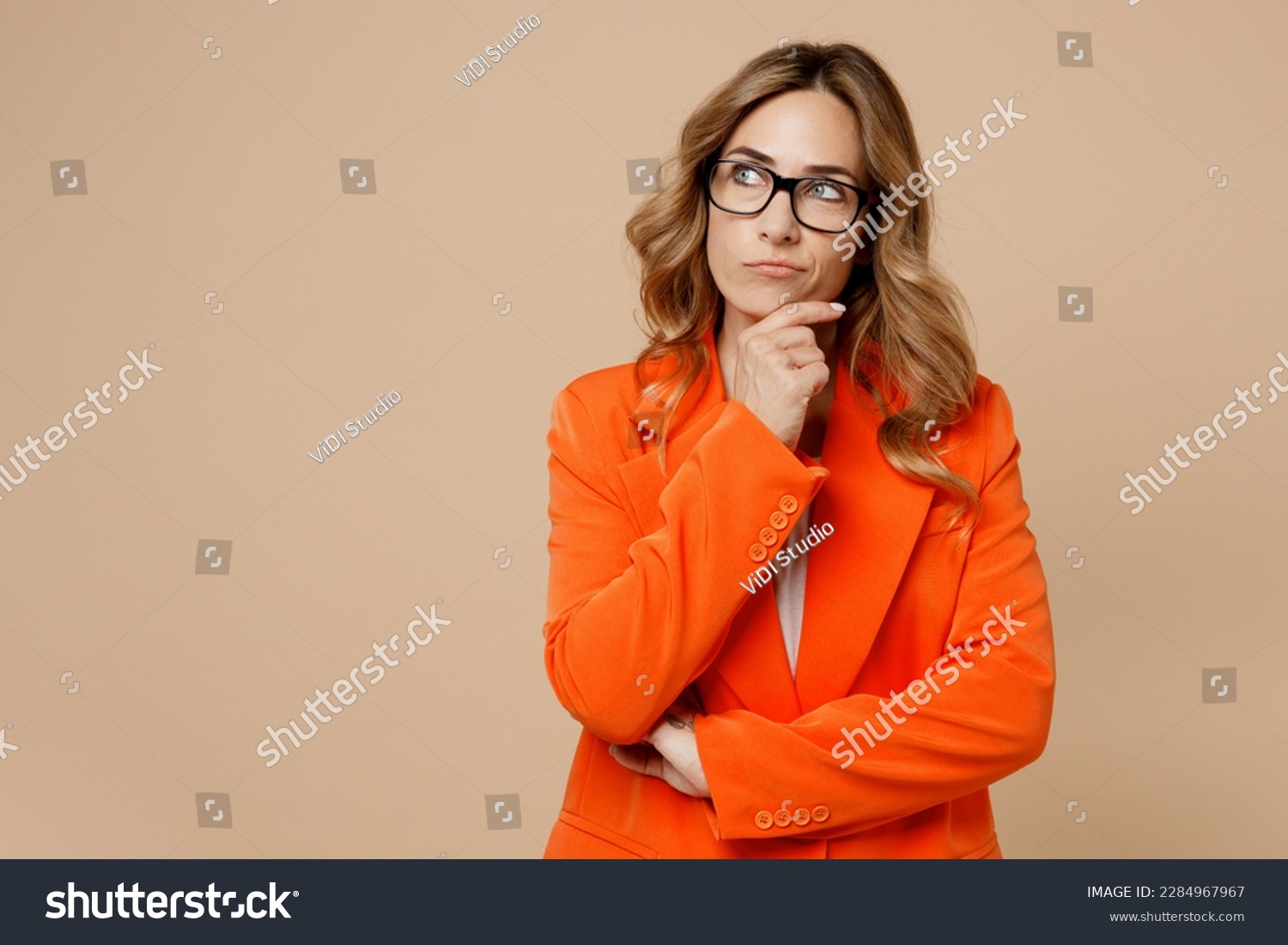 Young successful employee business woman corporate lawyer 30s wears classic formal orange suit glasses work in office prop up chin look aside on area isolated on plain beige color background studio #2284967967