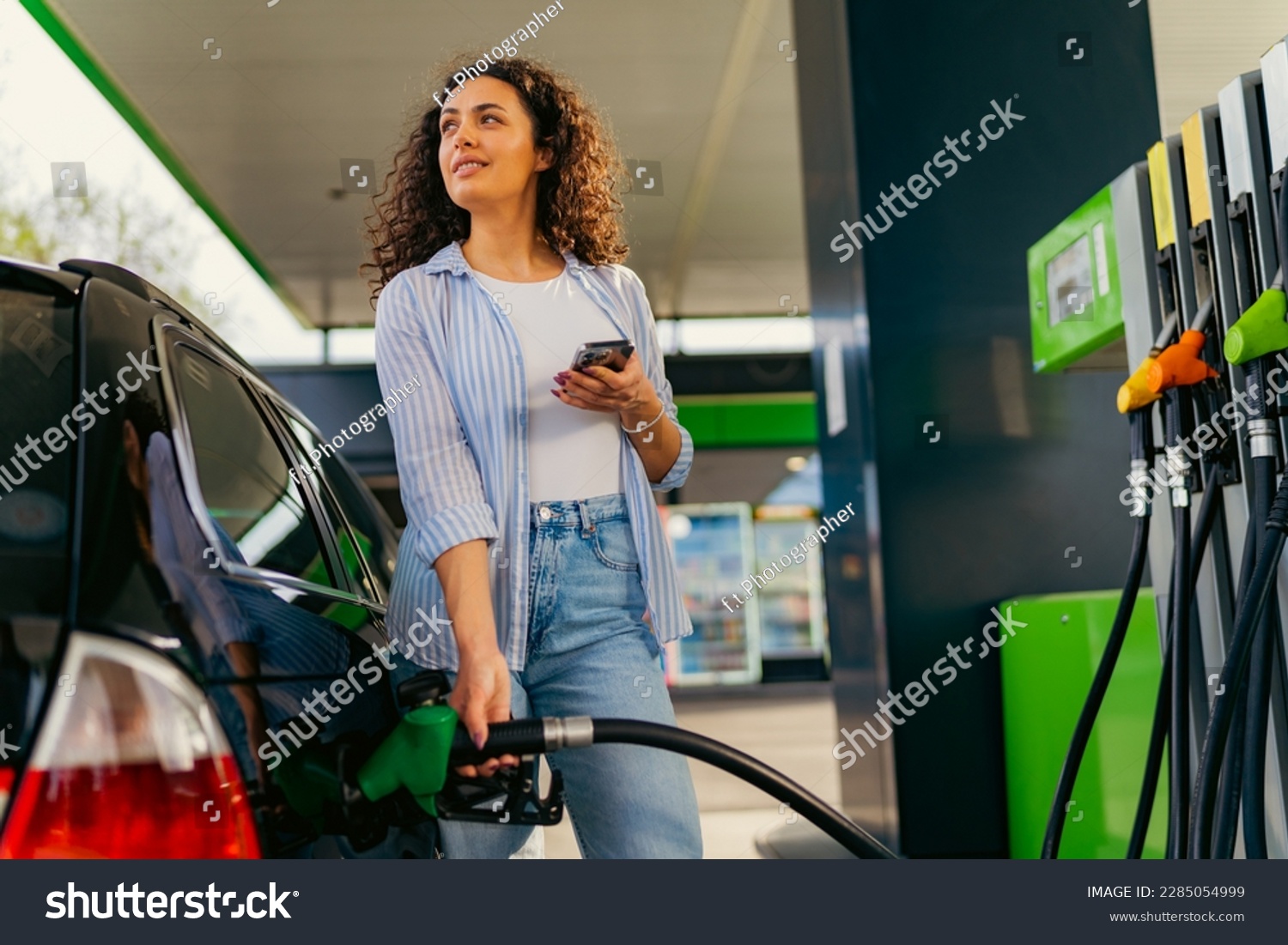 A young beautiful woman does not follow the safety rules and uses the phone while filling her car gas tank #2285054999