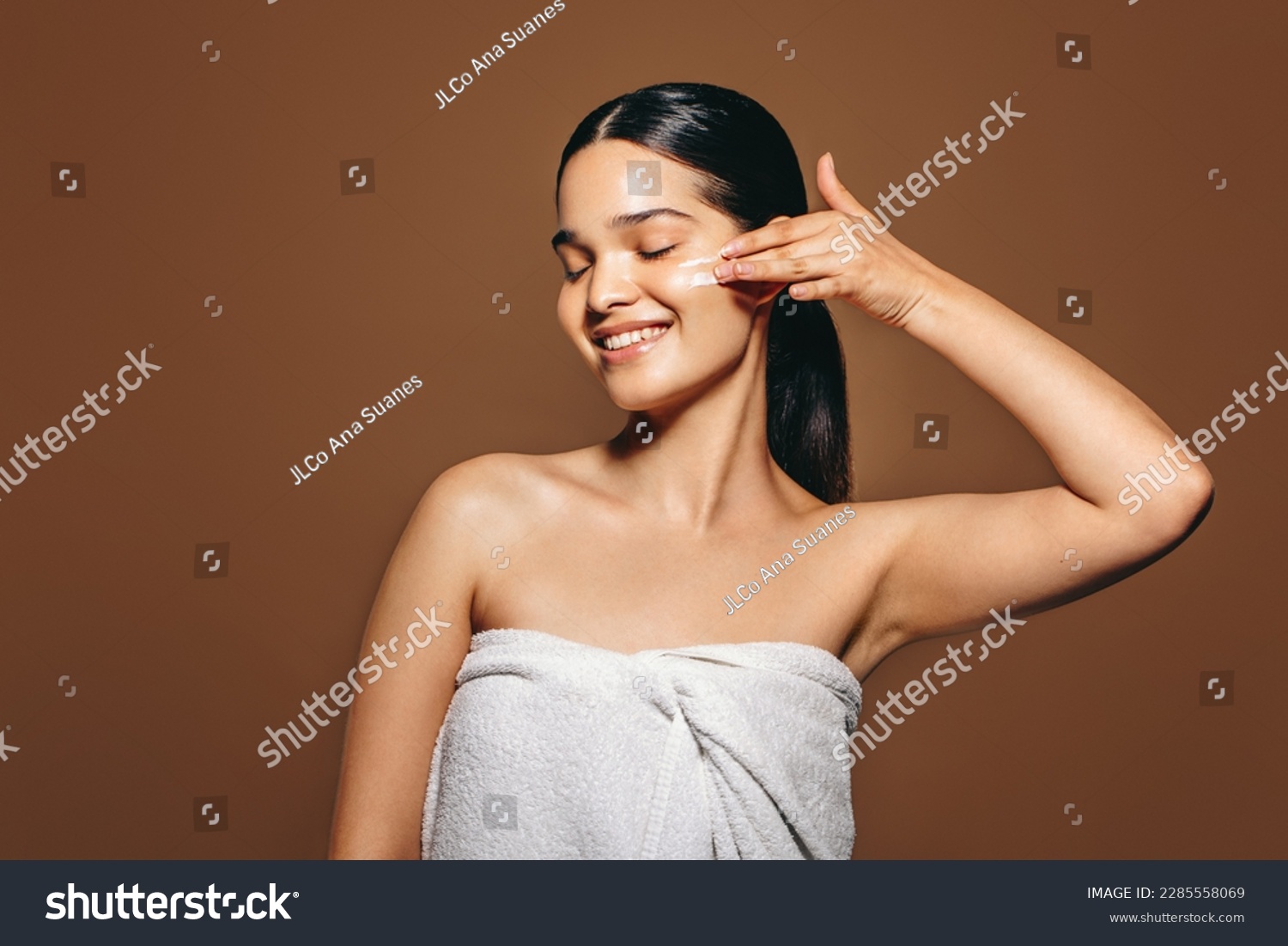 Female applying facial cream in a studio. Beautiful Hispanic woman pampering her skin using moisturizing lotion, enjoying practicing a healthy skincare routine for a radiant complexion. #2285558069