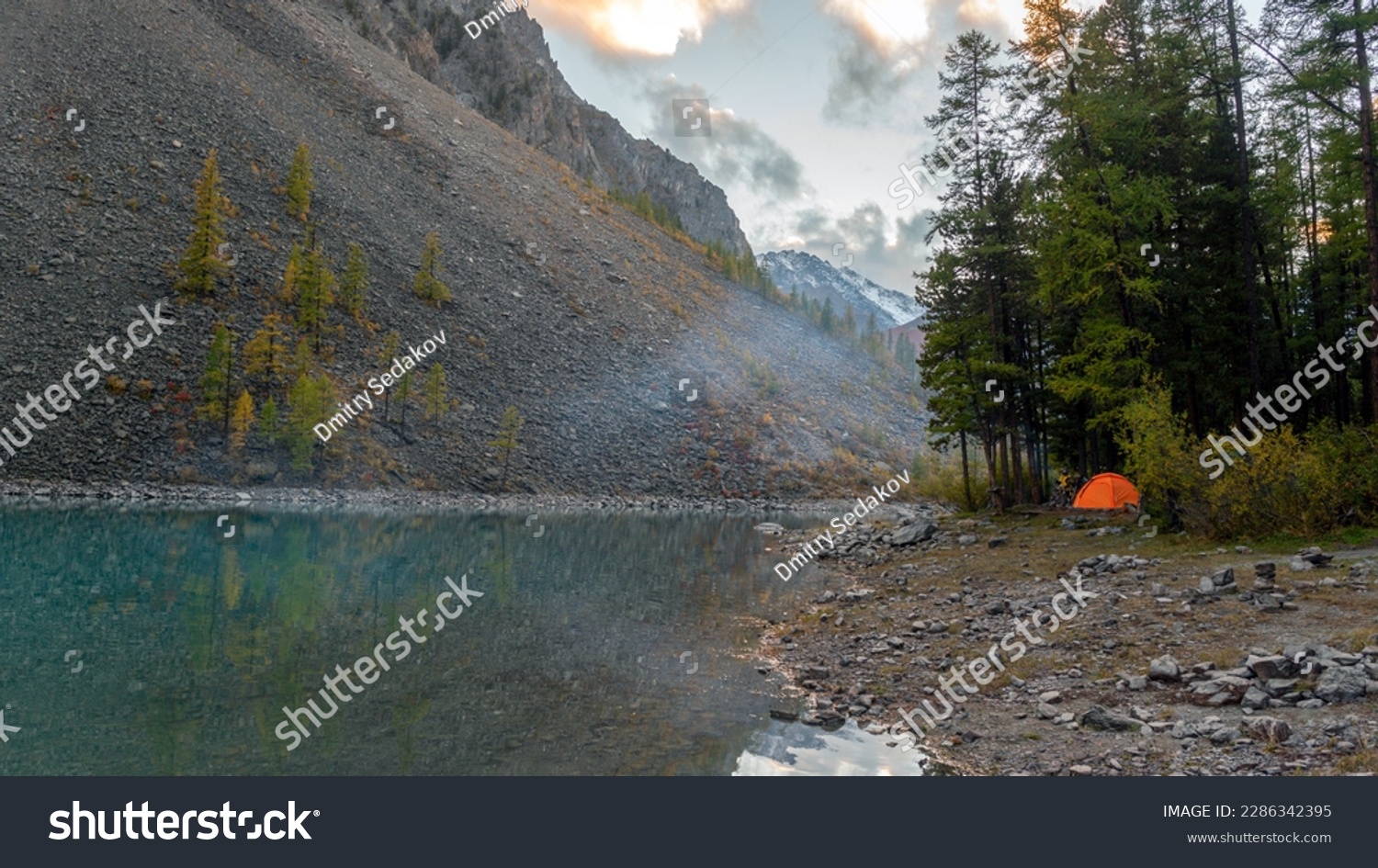 An orange tourist tent stands on the shore of the alpine lake Shavlinskoye near stone cliffs with smoke from a fire in Altai during the day. #2286342395