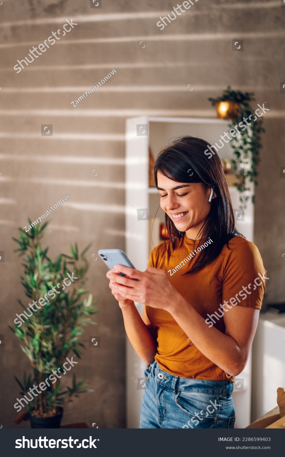 Beautiful woman checking social media while using smartphone and airpods at home. Smiling young female using mobile phone app and playing game, shopping online or reading news. Copy space. #2286599403