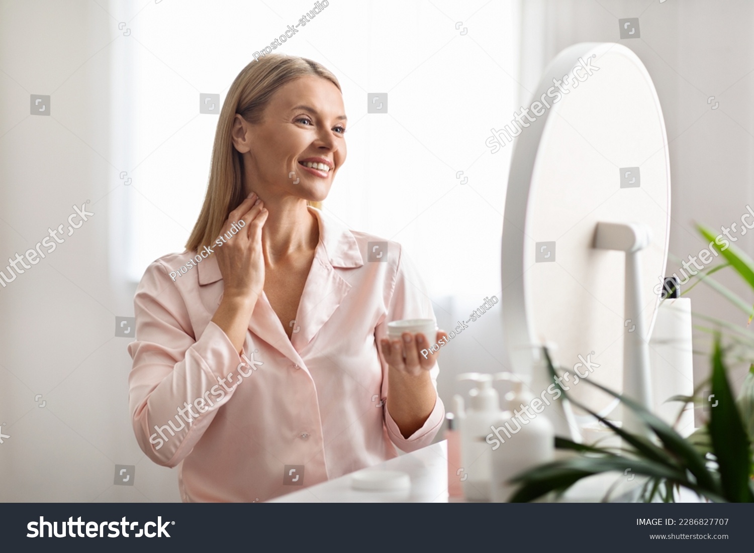 Beautiful middle aged woman applying moisturizing cream on her neck while sitting near mirror at home, attractive mature female enjoying domestic beauty routine, holding jar with nourishing lotion #2286827707
