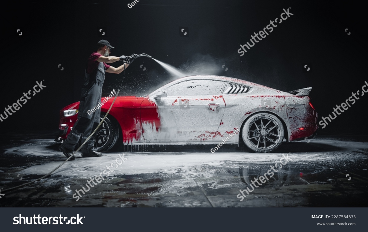 Car Wash Expert Using Water Pressure Washer to Clean a Red Modern Sportscar. Adult Man Washing Away Dirt, Preparing an American Muscle Car for Detailing. Creative Cinematic Photo with Luxury Vehicle #2287564633