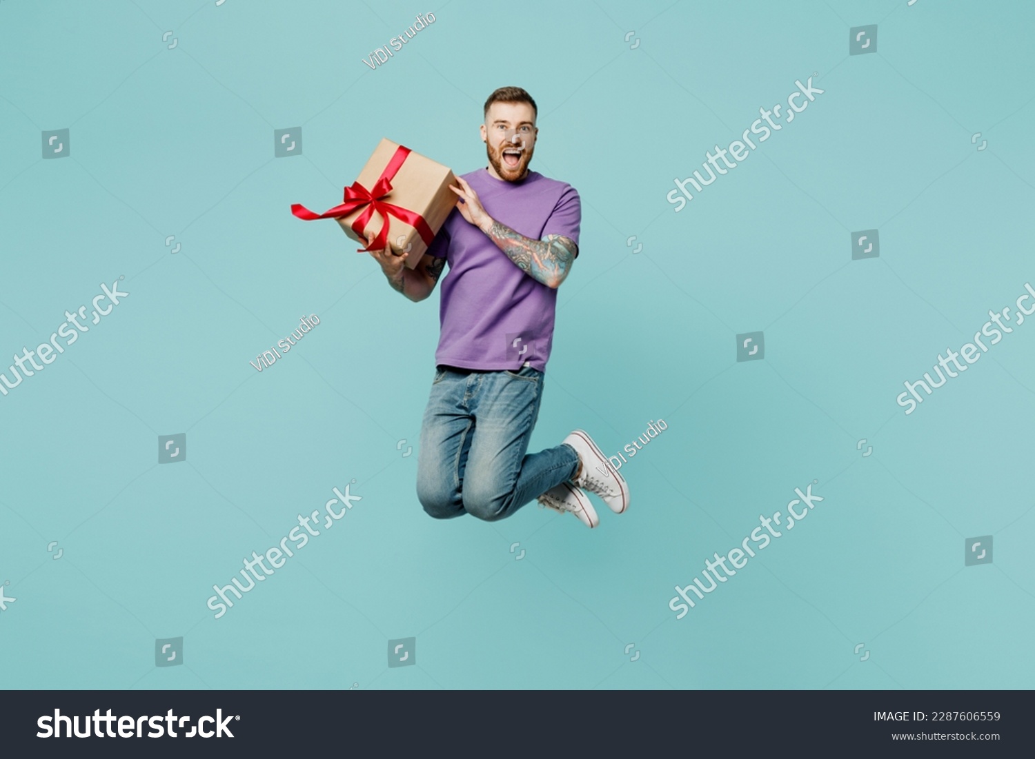 Full body excited fun young man he wears purple t-shirt jump high hold in hand present box with gift ribbon bow isolated on plain pastel light blue cyan background studio portrait. Lifestyle concept #2287606559