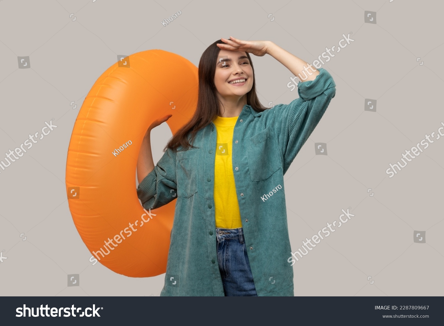 Satisfied woman looking far with hand near eyes and holding orange rubber ring, waiting to travel to tropical country, wearing casual style jacket. Indoor studio shot isolated on gray background. #2287809667