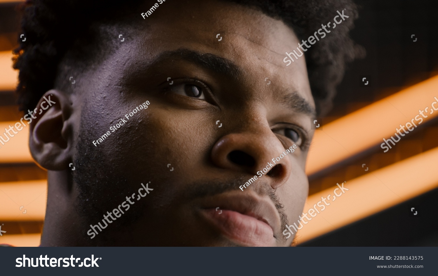 Young African American man straight and serious face with confident deep look. Focused worker looks away, blinks eyes, poses on camera. LED lamps on background in studio or office. Close up portrait. #2288143575