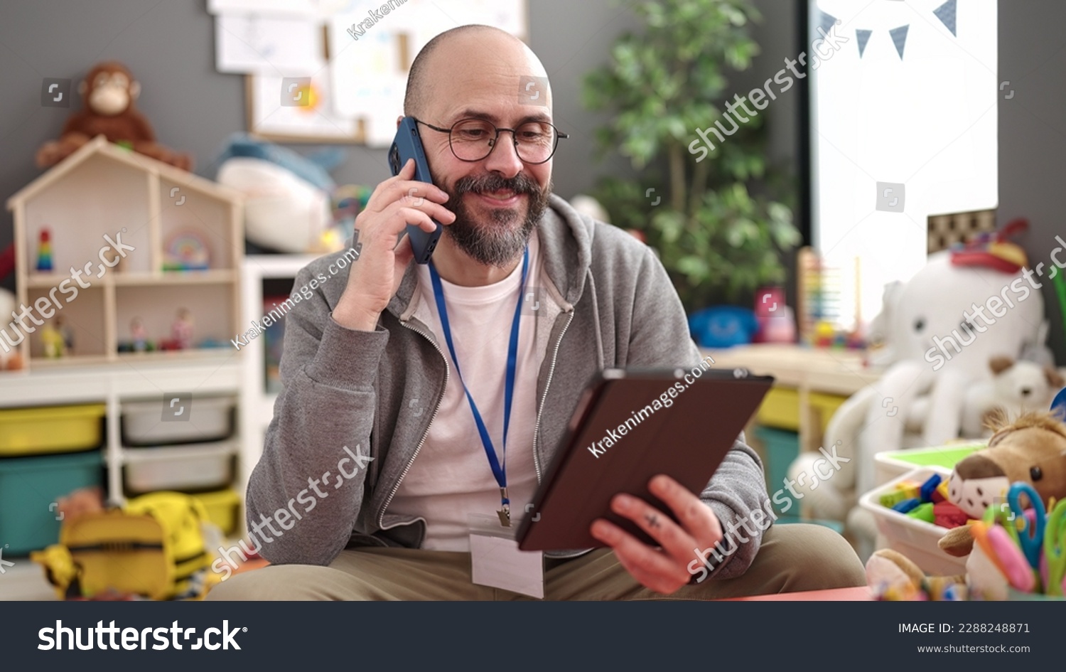 Young bald man preschool teacher using touchpad speaking on the phone at kindergarten #2288248871