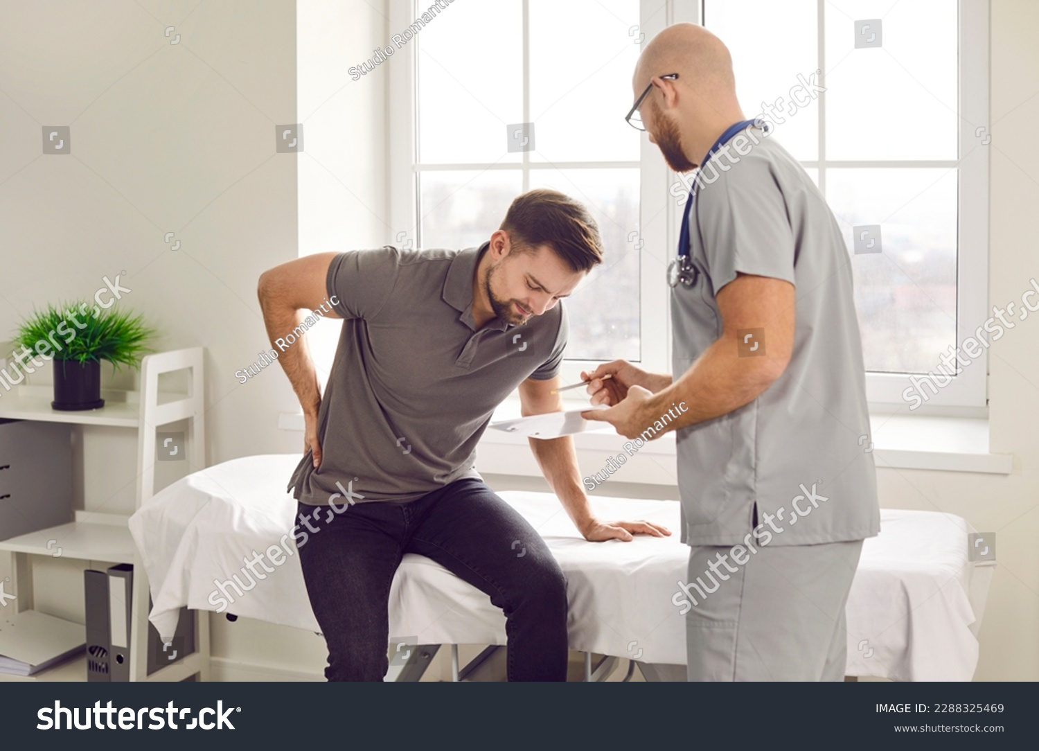 Patient with lower back pain comes to the doctor. Young man suffering from lumbar pain sitting on a medical bed at the clinic and asking a professional physician to help him #2288325469