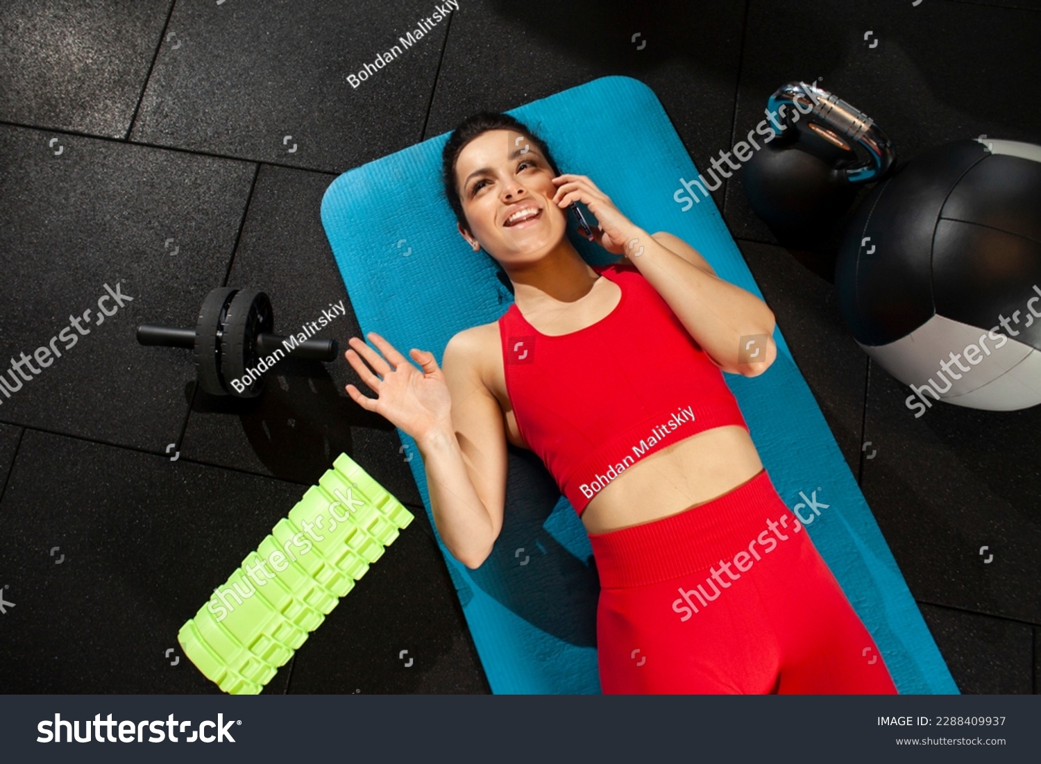 athletic woman in red sportswear lies on yoga matte and talks on the phone in black gym, girl makes phone call in fitness room, attractive woman in fitness club communicates #2288409937
