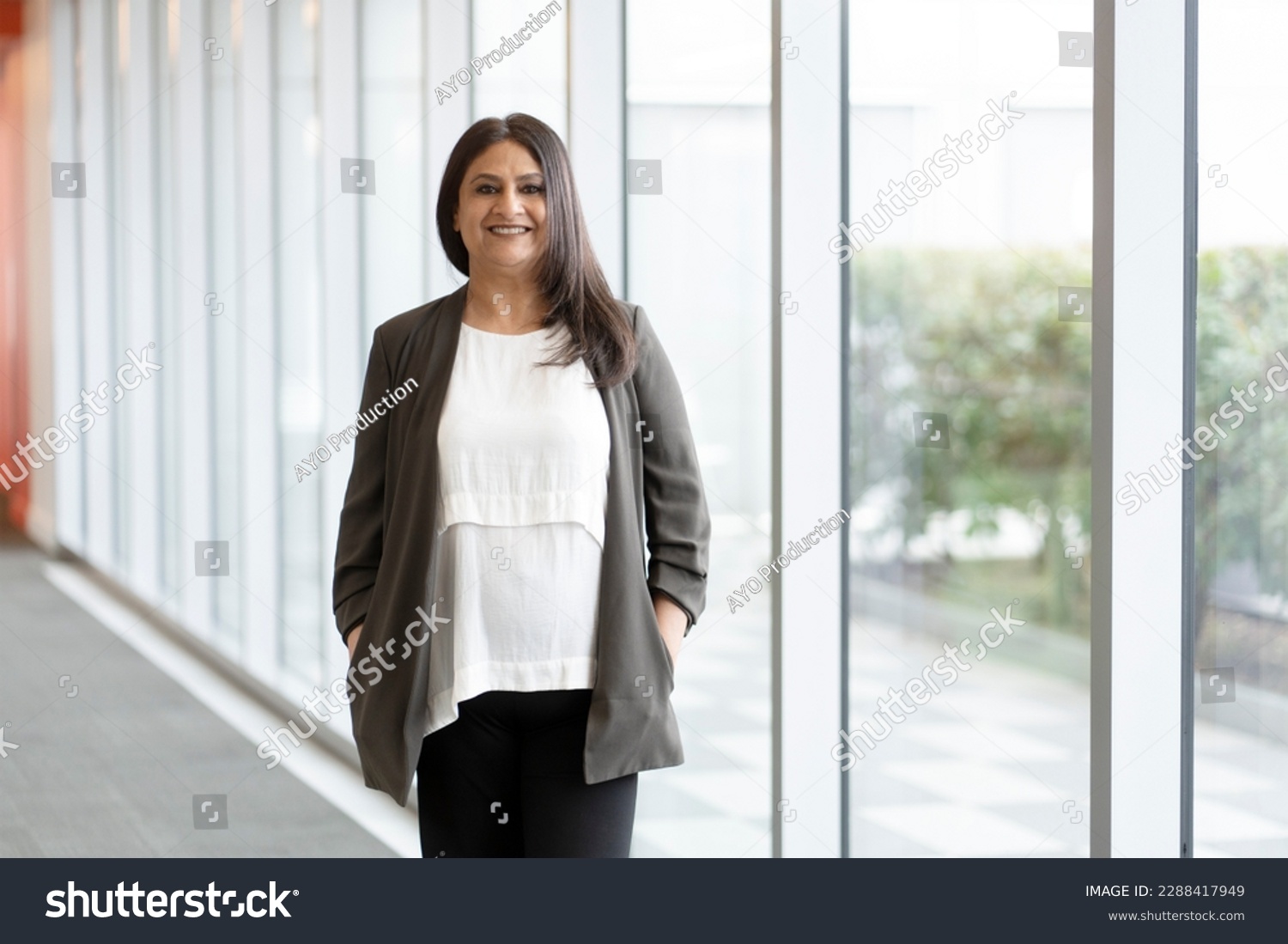 Portrait of confident mature Indian woman standing by large windows in the corporate office, smiling cutely looking at camera. Successful Asian people and business concept #2288417949