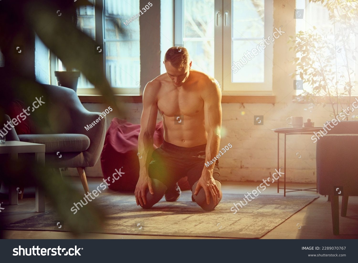 Young man with strong, muscular, relief body posing shirtless, doing home training on daytime with sunlight. Breathing. Concept of sportive lifestyle, body and health care, fitness, health #2289070767