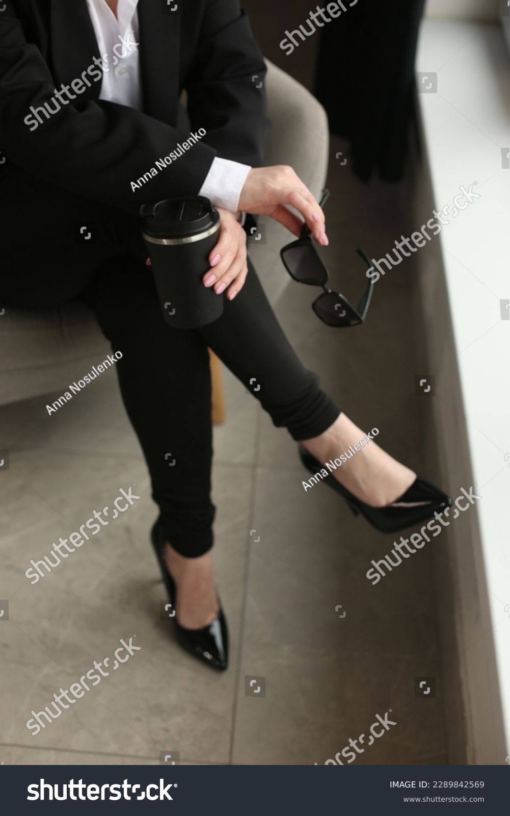 a girl sitting on an armchair in a black suit with a white blouse on her feet high-heeled shoes and leggings in her hands a thermos mug and glasses #2289842569