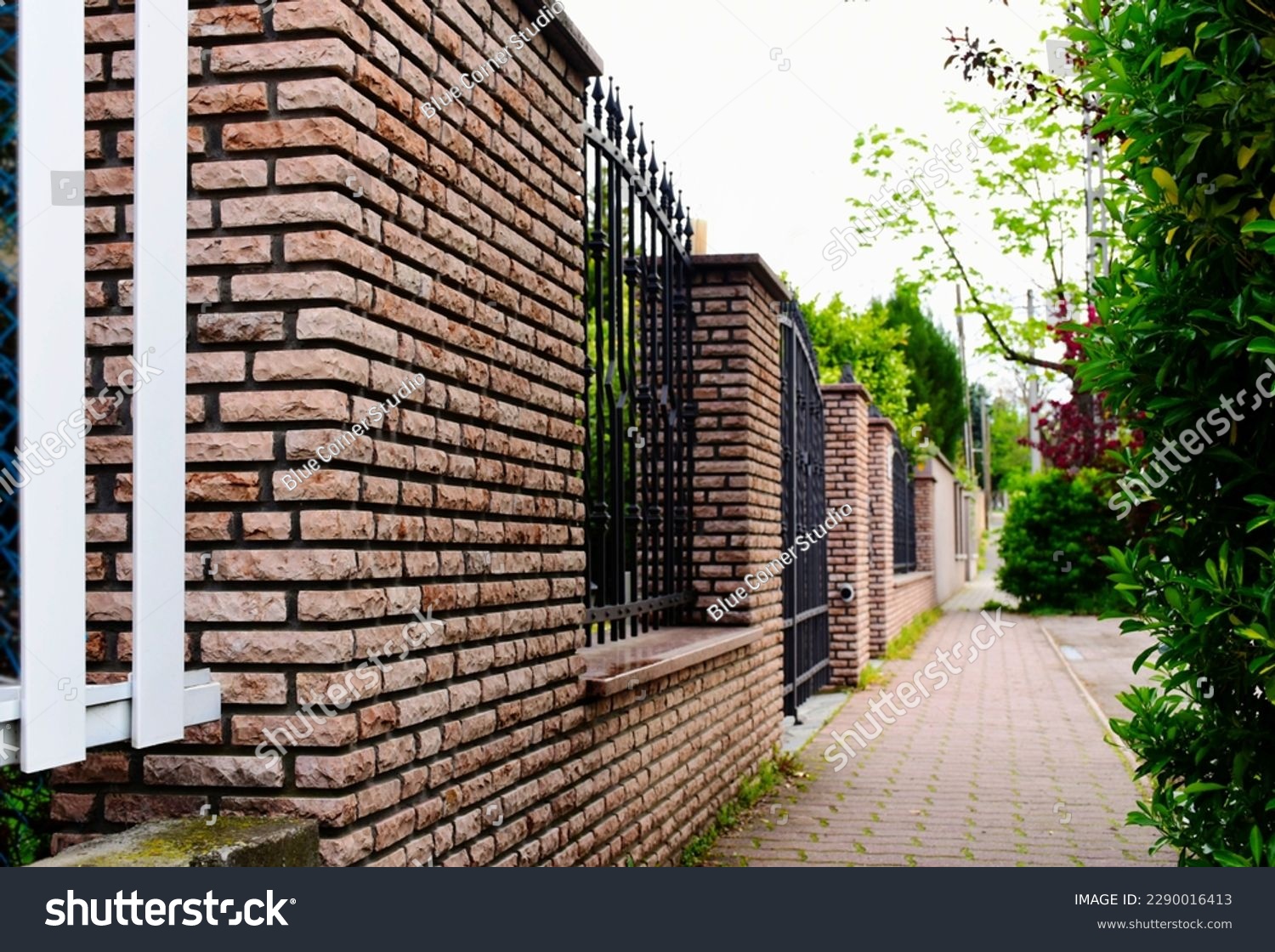 decorative wrought iron picket fence panels and stone fence piers. green garden, lush trees, plants and foliage. home ownership concept. property protection. concrete pavement. diminishing perspective #2290016413