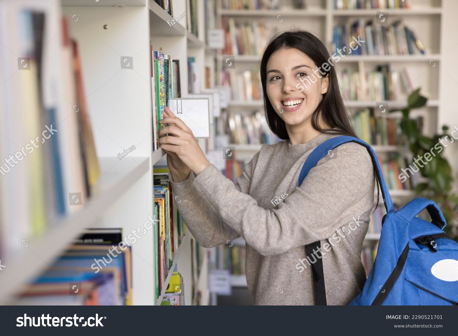 Pretty girl chooses books, standing in public library. Caucasian female student prepares for university admission looks interested and motivated, take textbook staring aside. Education, new knowledge #2290521701