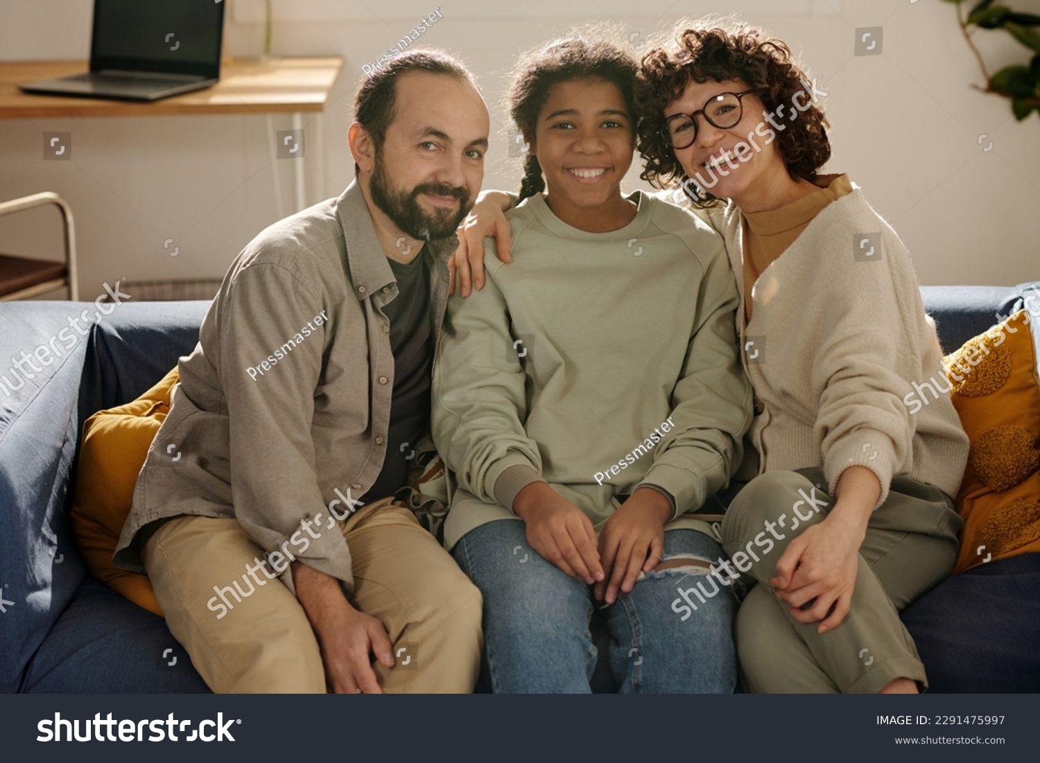 Portrait of happy parents sitting together with their adopted daughter on sofa and smiling at camera #2291475997