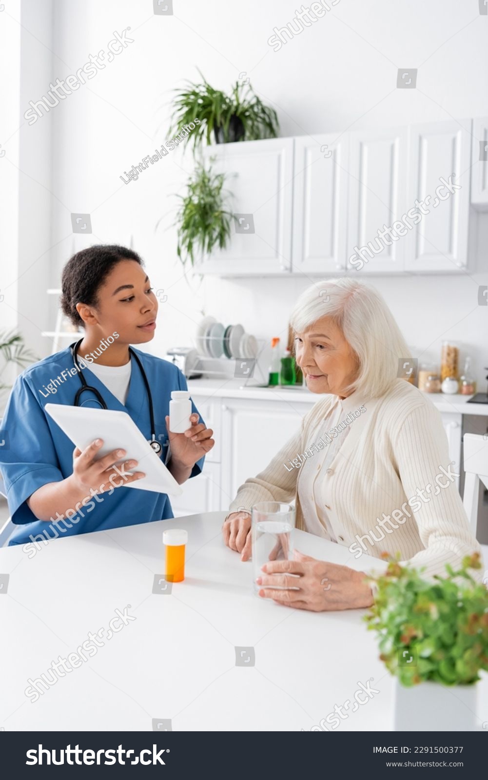 brunette multiracial nurse using digital tablet and holding medication while talking to retired woman with grey hair #2291500377