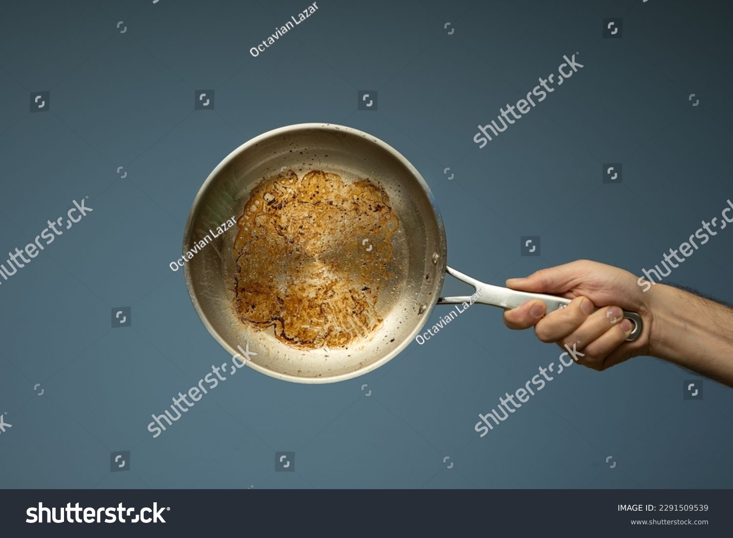 Dirty oily burnt metal frying pan held in hand by a male hand. Close up studio shot, isolated on a light blue background. #2291509539