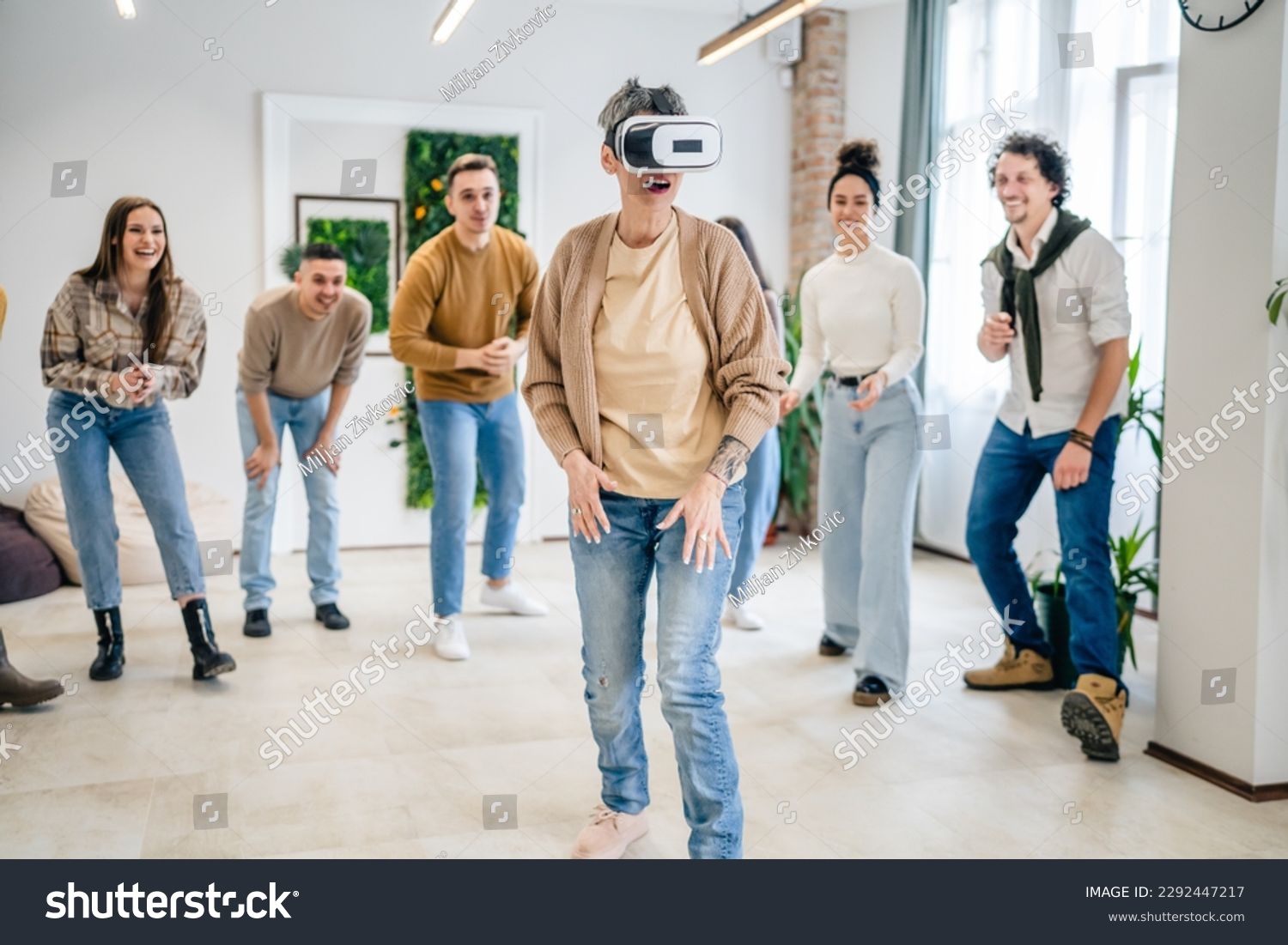 One woman mature senior caucasian female in front of group of men and women friends enjoy virtual reality VR headset at work having fun together during team building seminar real people bright filter #2292447217