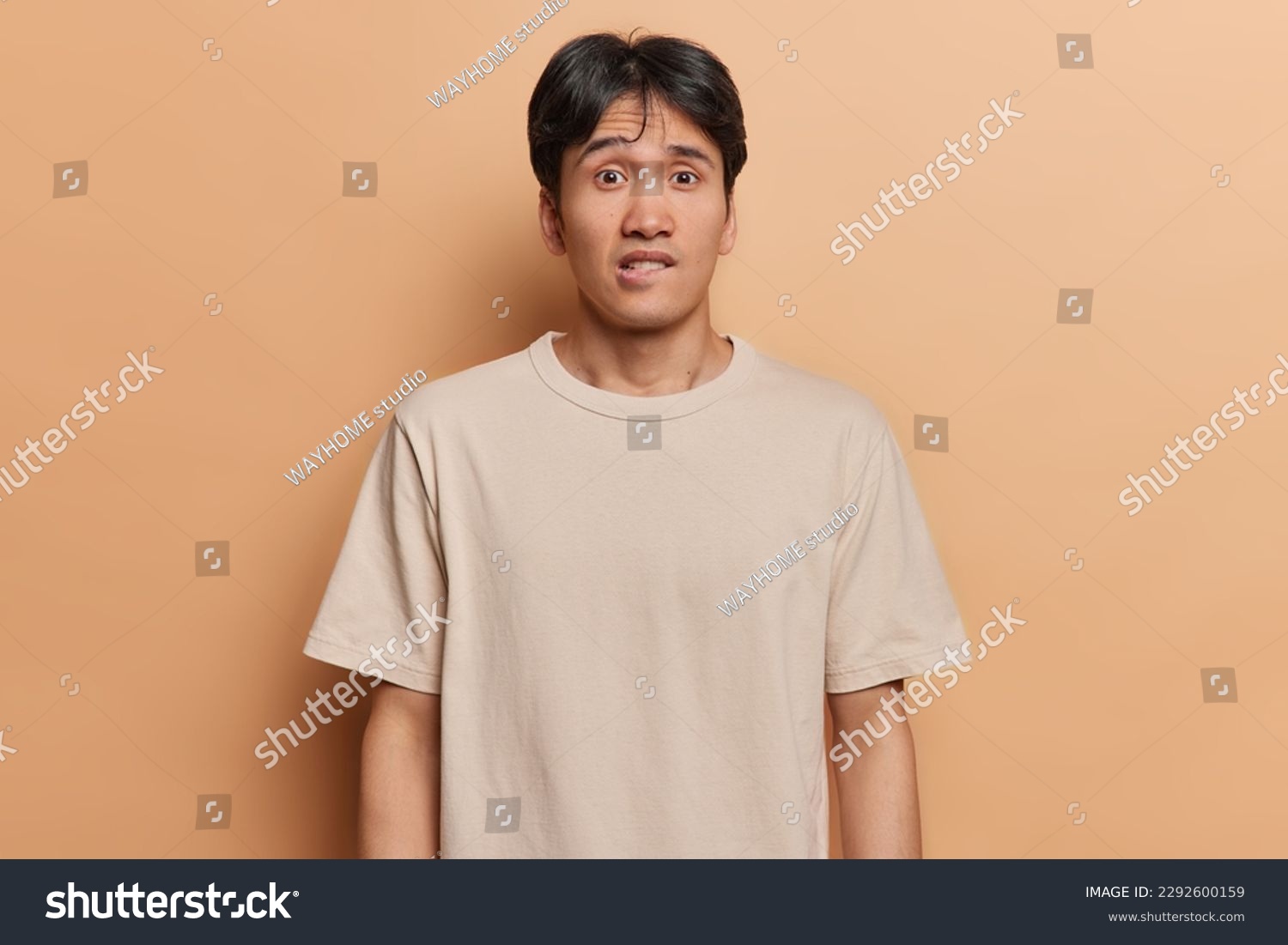 Waist up shot of worried Japanese man bites lips looks anxious and surprisingly at camera doesnt know way out from difficult situation dressed in casual t shirt isolated over brown background #2292600159