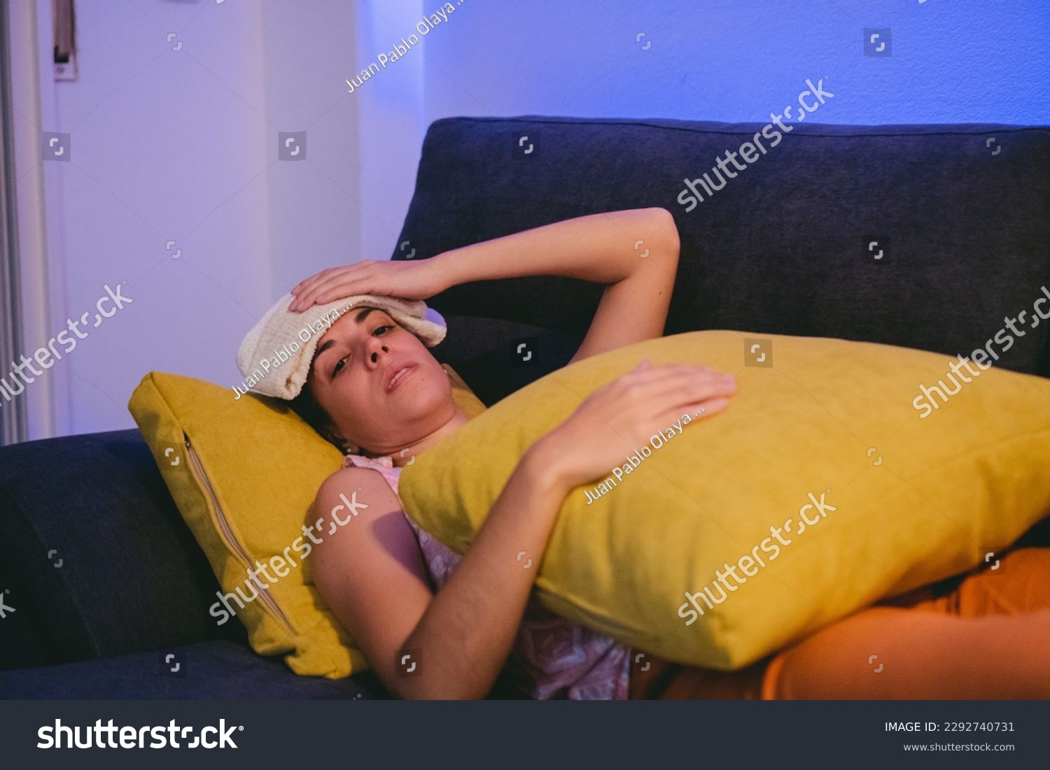 Sick young Latin woman lying on a sofa with a towel on her head to bring down her fever while hugging a cushion. #2292740731