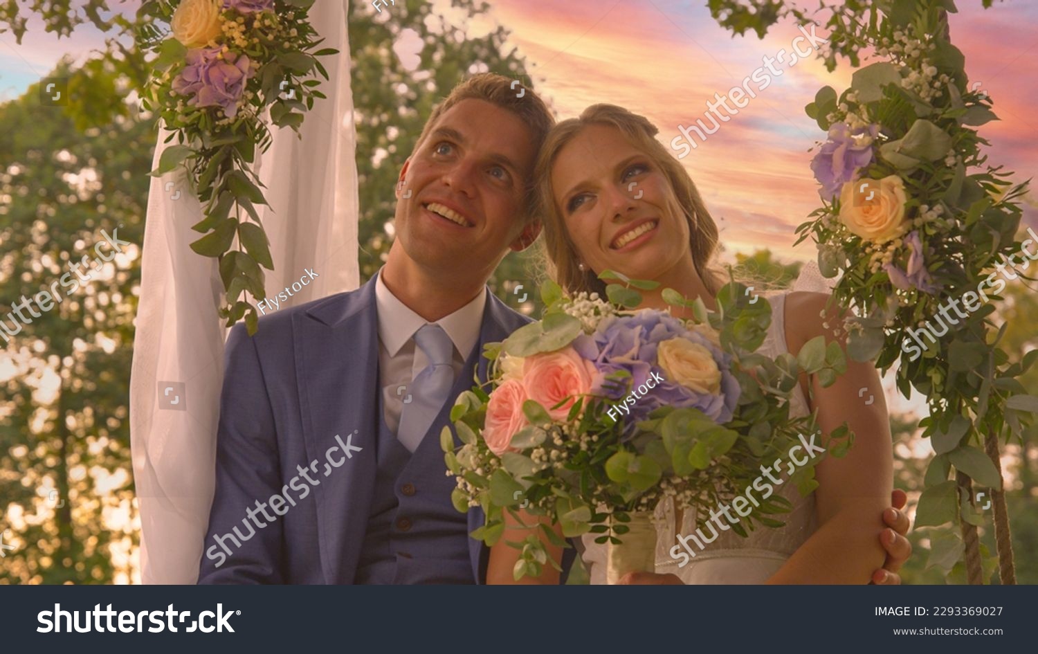 PORTRAIT, LENS FLARE: Smiling newlyweds posing on a beautifully decorated swing. Lovely bride and handsome groom in embrace of each other under colorful summer sunset clouds on their wedding day. #2293369027
