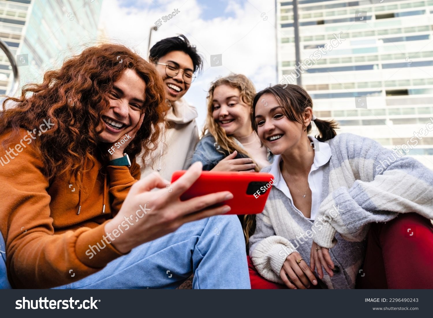 Group of young gen z friends sitting together in the city using cell phone app to share funny content #2296490243