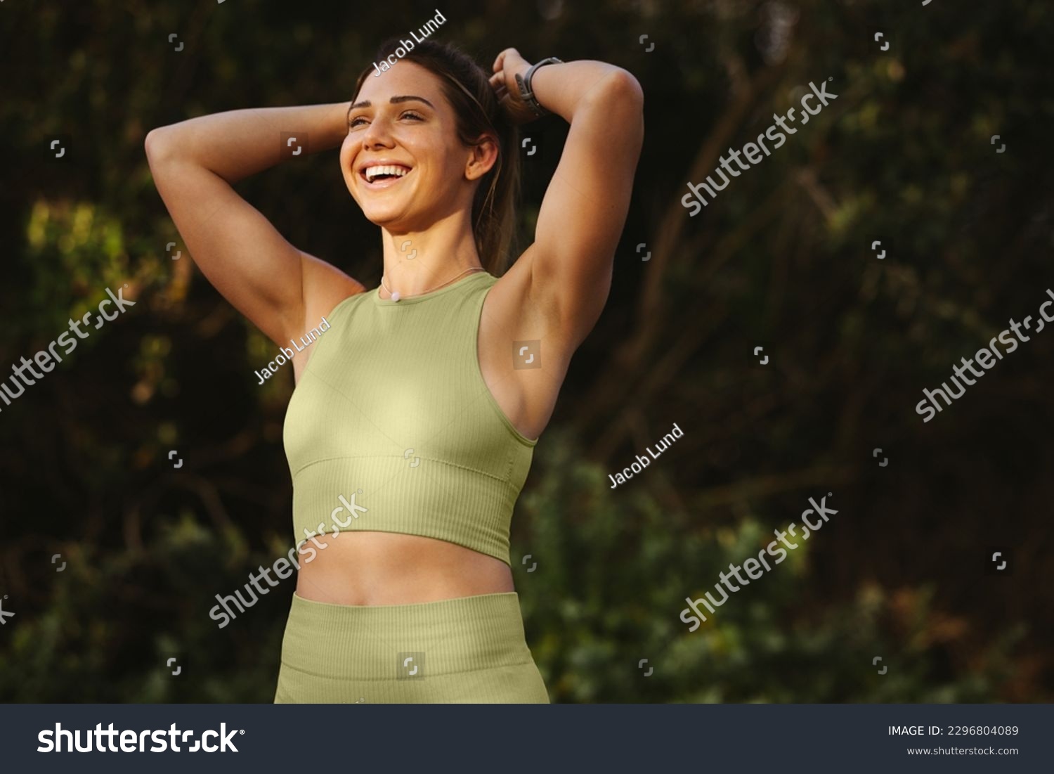 Woman warming up for a morning workout outdoors. Happy sports woman tying her hair and preparing for yoga. Fit woman standing against a nature background. #2296804089