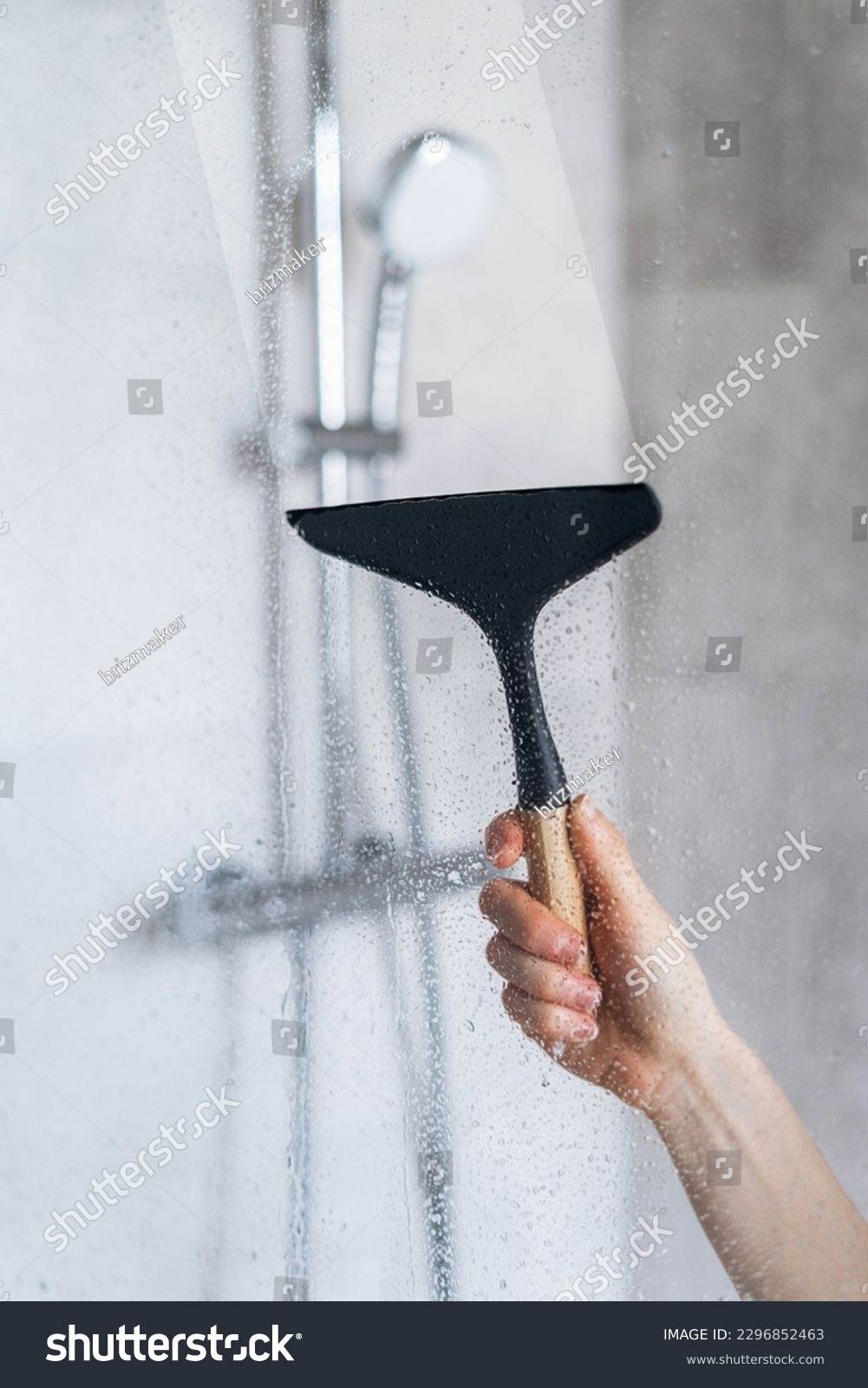 Closeup view of woman hand holding glass wiper and cleaning dirt and drops on shower partition in the bathroom. View on shower system as background. Cleaning service, washing concept #2296852463