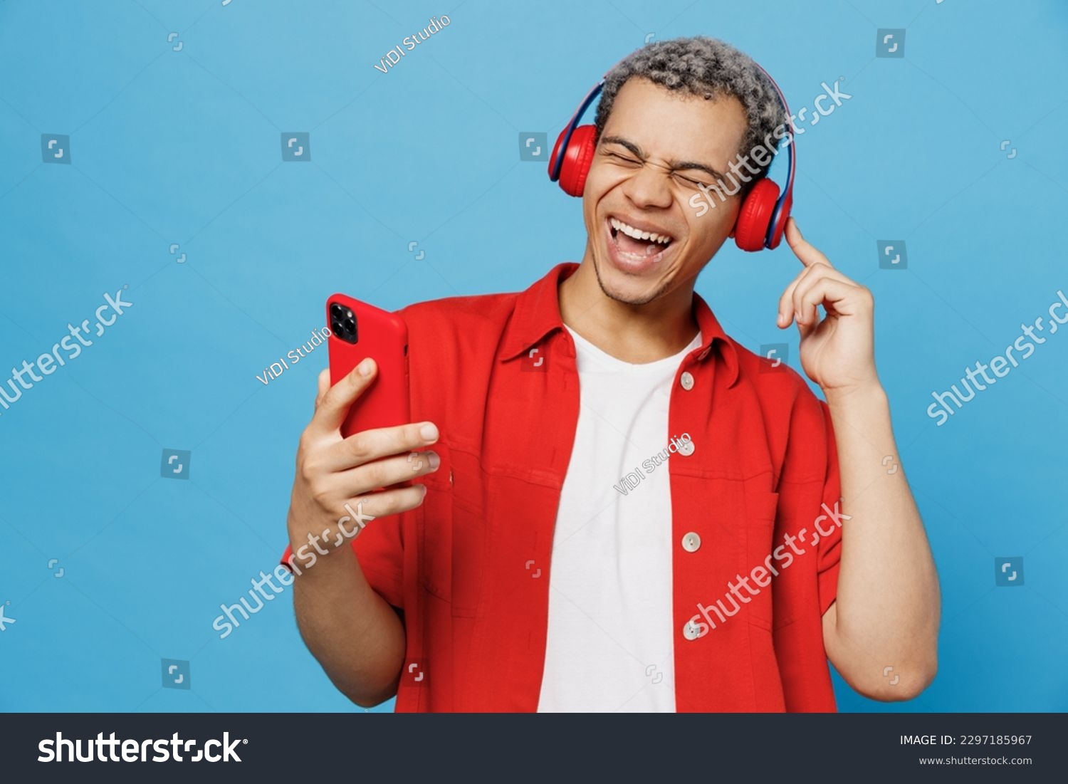 Young cheerful happy fun vivid man of African American ethnicity 20s he wearing red shirt headphones listen music sing song use mobile cell phone isolated on plain pastel light blue cyan background #2297185967