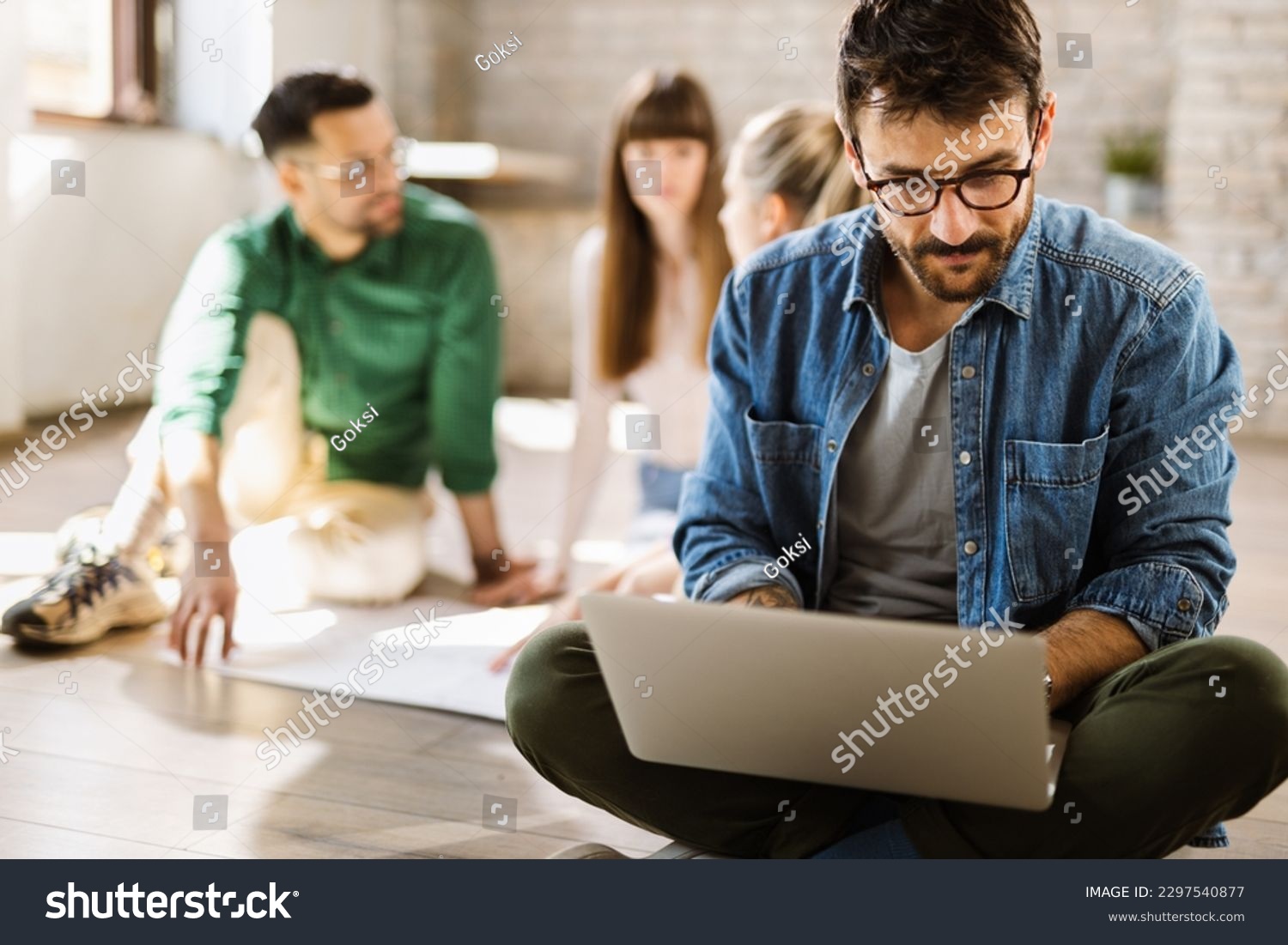 Young enterpreneur using computer while sitting on the floor in the office. There are people in the background #2297540877