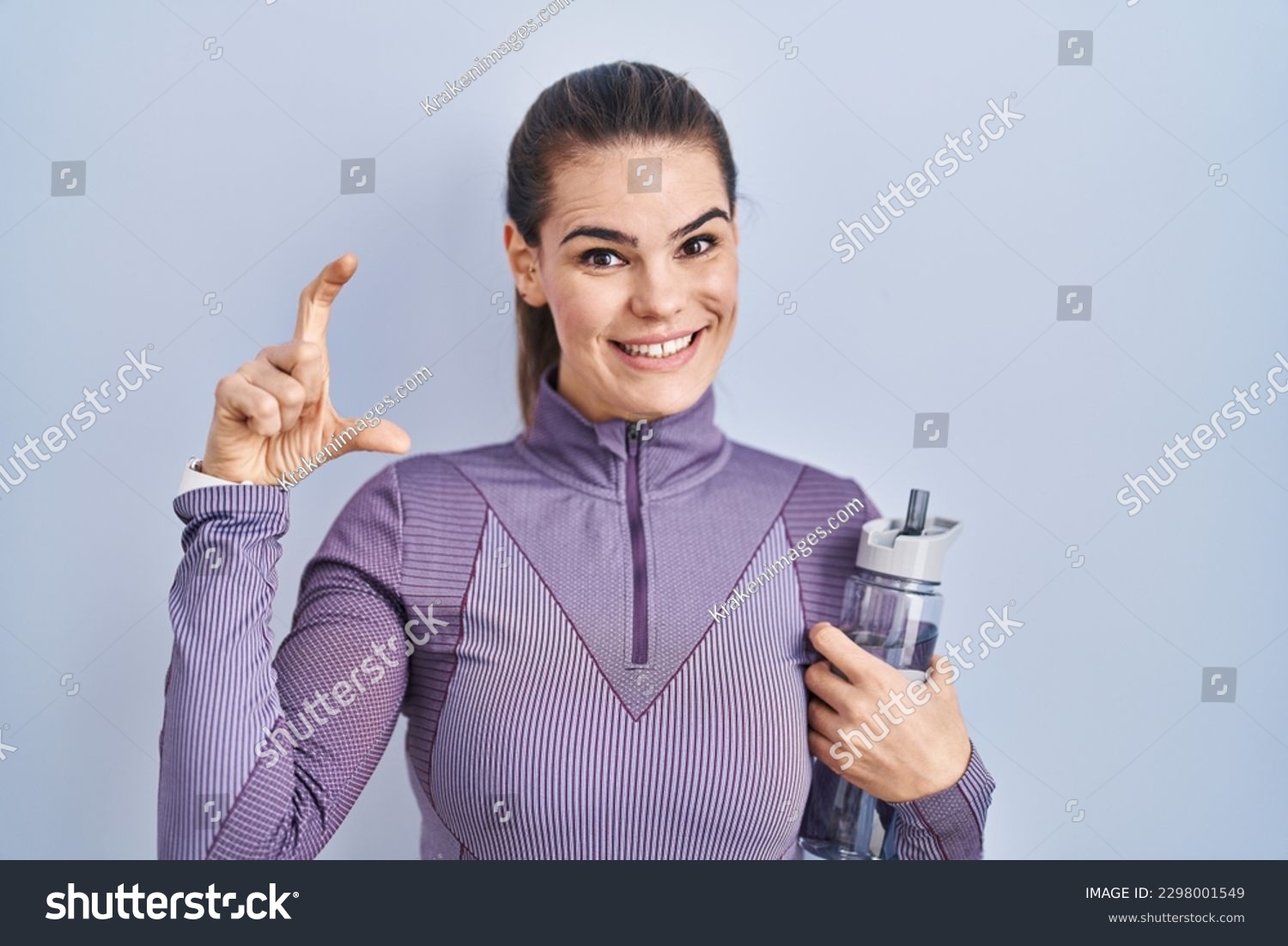 Beautiful woman wearing sportswear holding water bottle smiling and confident gesturing with hand doing small size sign with fingers looking and the camera. measure concept.  #2298001549