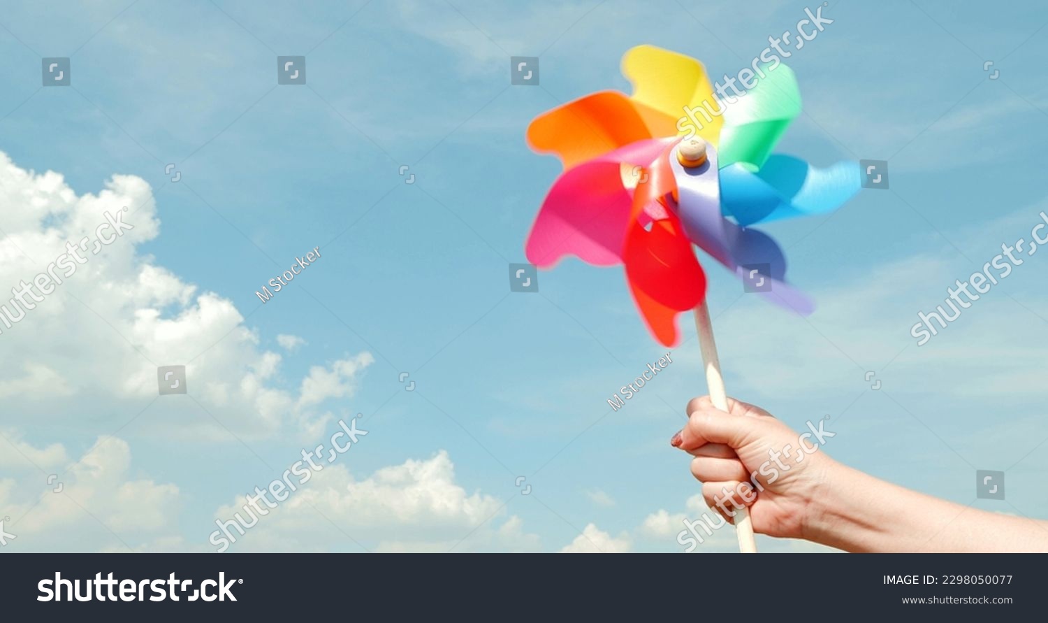Motion blurred of colorful pinwheel. Woman hand holding colorful plastic windmills on blue sky background. Green energy concept #2298050077