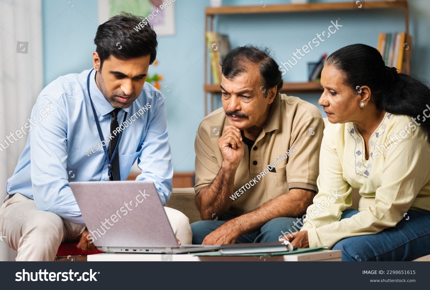 Indian banker explaining about insurance policy or savings scheme on laptop to senior couple while sitting on sofa at home - concept of banking support, and bonding #2298651615