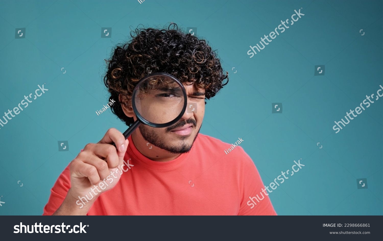 funny young latin spanish guy with a magnifying glass in his hands looks out or looks for something, dressed in casual, on a blue studio background. #2298666861