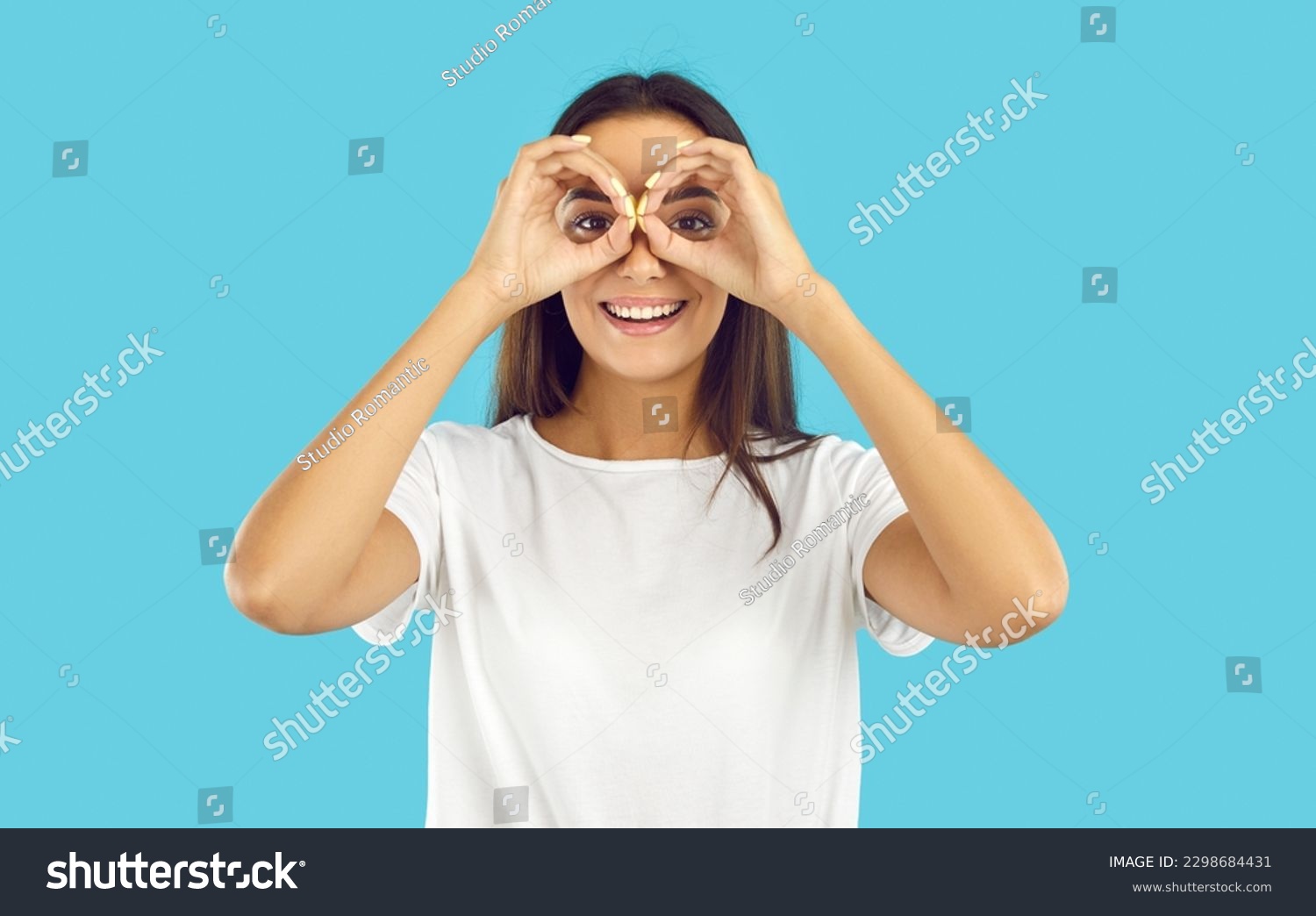Happy pretty woman looking through pretend binoculars. Cheerful beautiful young brunette lady in white T shirt forming circles with her hands, doing binoculars gesture, looking in distance and smiling #2298684431