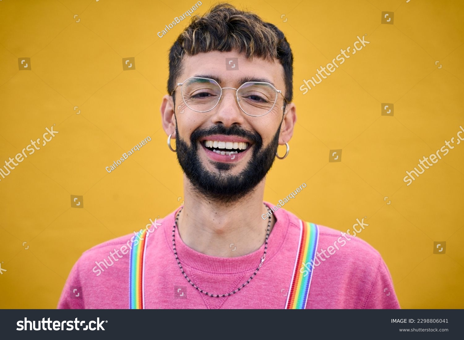 Close up smiling cheerful young Caucasian man holding his rainbow lgbt suspenders. Gay people posing for photo yellow background. Cheerful gay bearded person happy studio photo isolated. Millennials. #2298806041