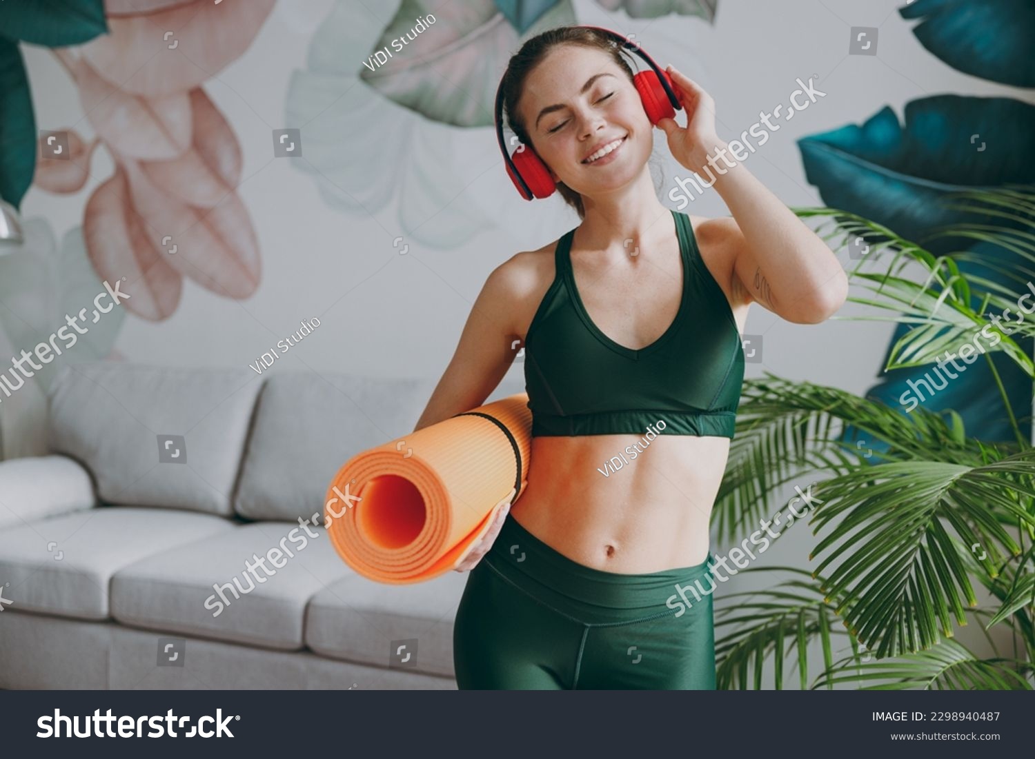 Young calm strong sporty athletic fitness trainer instructor woman wear green tracksuit headphones listen music hold in hand yoga mat training do exercises at home gym indoor. Workout sport concept #2298940487