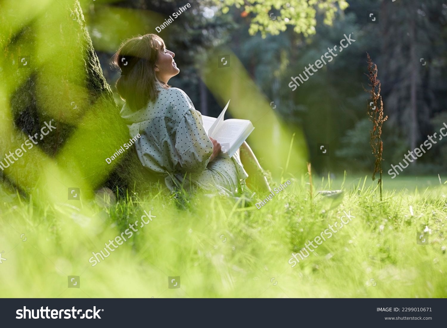 A young woman sits in a park leaning against a tree, reading a book in spring or summer, relaxed and happy, enjoying the sun. #2299010671
