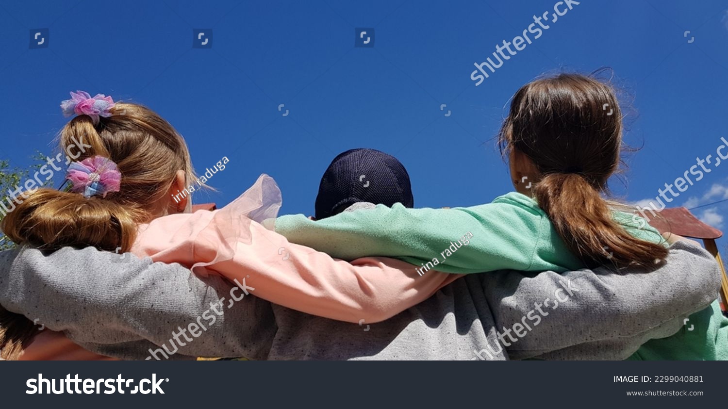 Three teenagers: two girls and a boy between them, hugging their shoulders, in the sun in the blue sky in spring (macro, rear view from the back). #2299040881