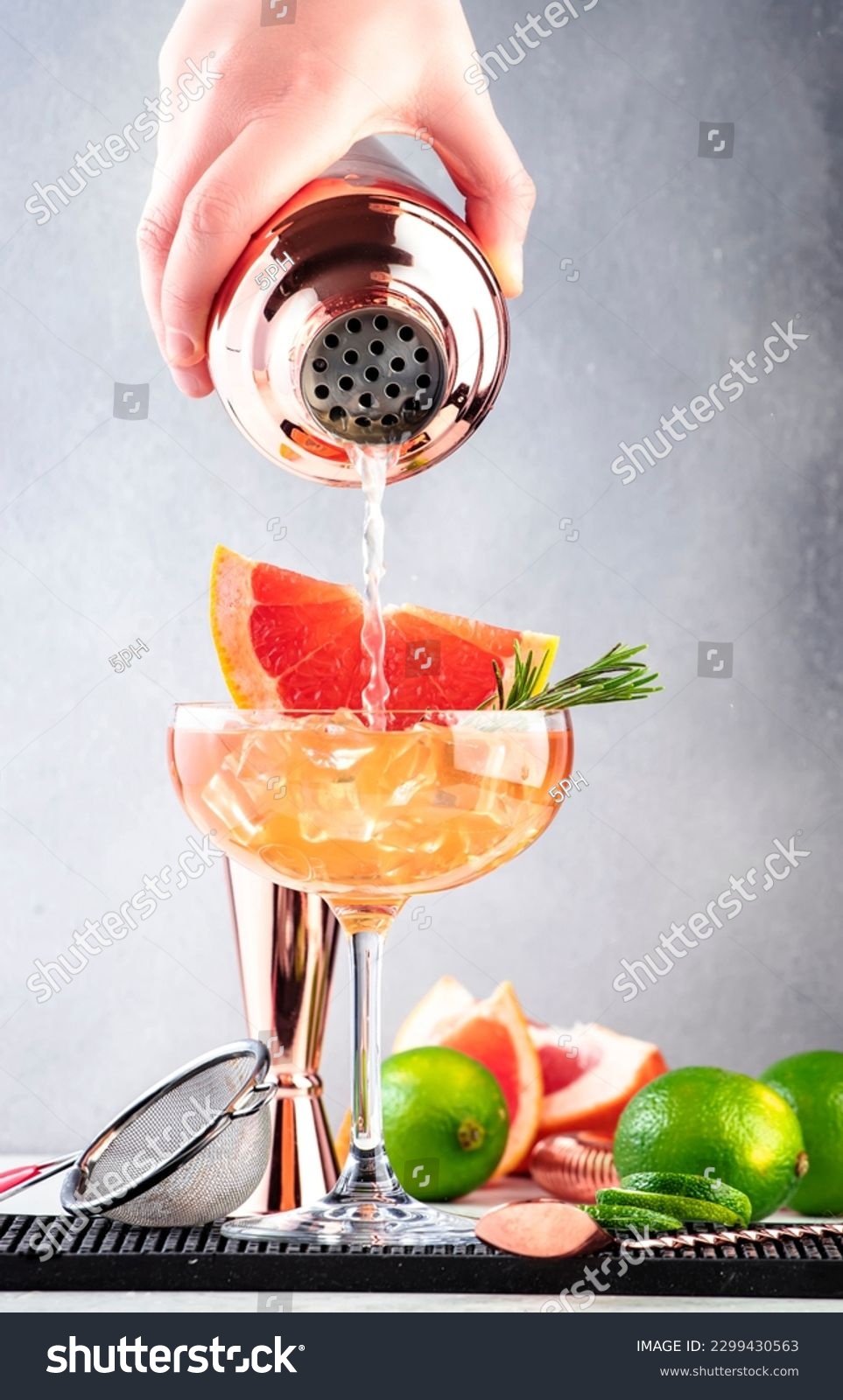 Bartender pours cocktail from shaker into glass. Grapefruit daiquiri alcoholic cocktail with white rum, syrup, fruit juice, lime and ice in garnished margarita glass. Gray background, bar tools #2299430563