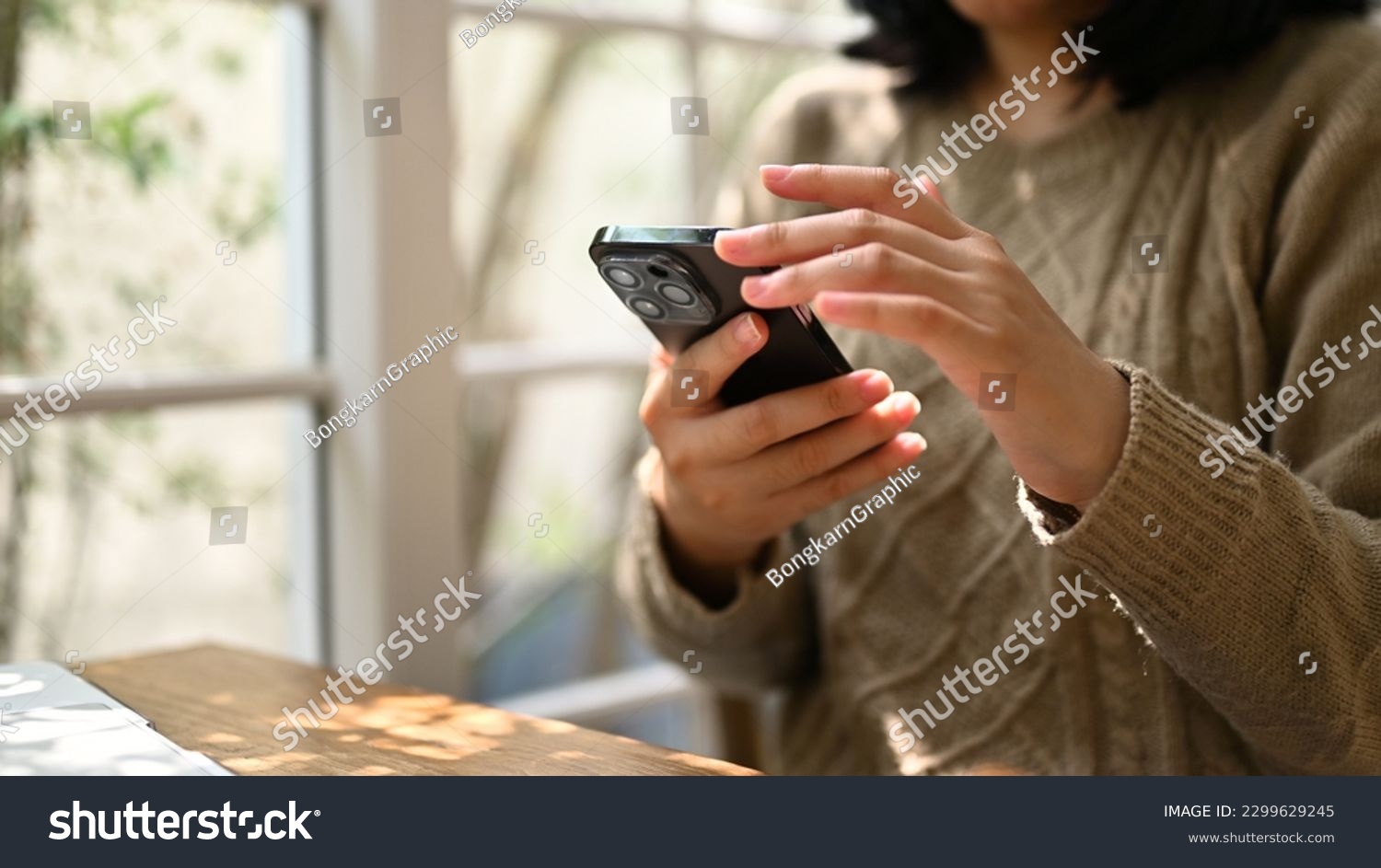 Close-up shot of an Asian woman in cozy sweater using her smartphone, chatting with someone or scrolling on her phone while relaxing at a cafe. #2299629245