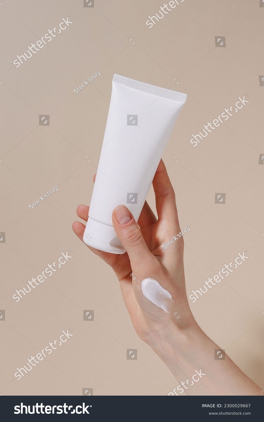Vertical image of a female hand with a white smear holding a blank tube of moisturizing cream on a beige isolated background. Hand care, natural cosmetics and beauty concept. Image for your design #2300029667