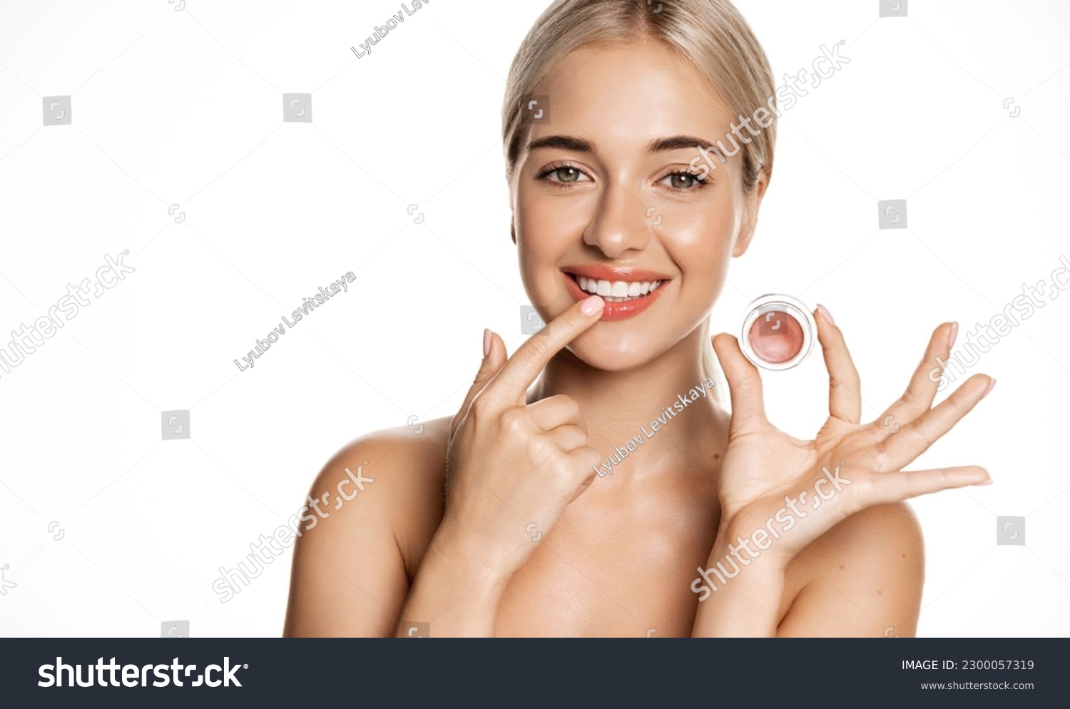 Close up of blond smiling beauty model, girl applies lip balm, pink gloss for natural shine and glow, uses makeup on clean, perfect skin without blemishes, white background. #2300057319