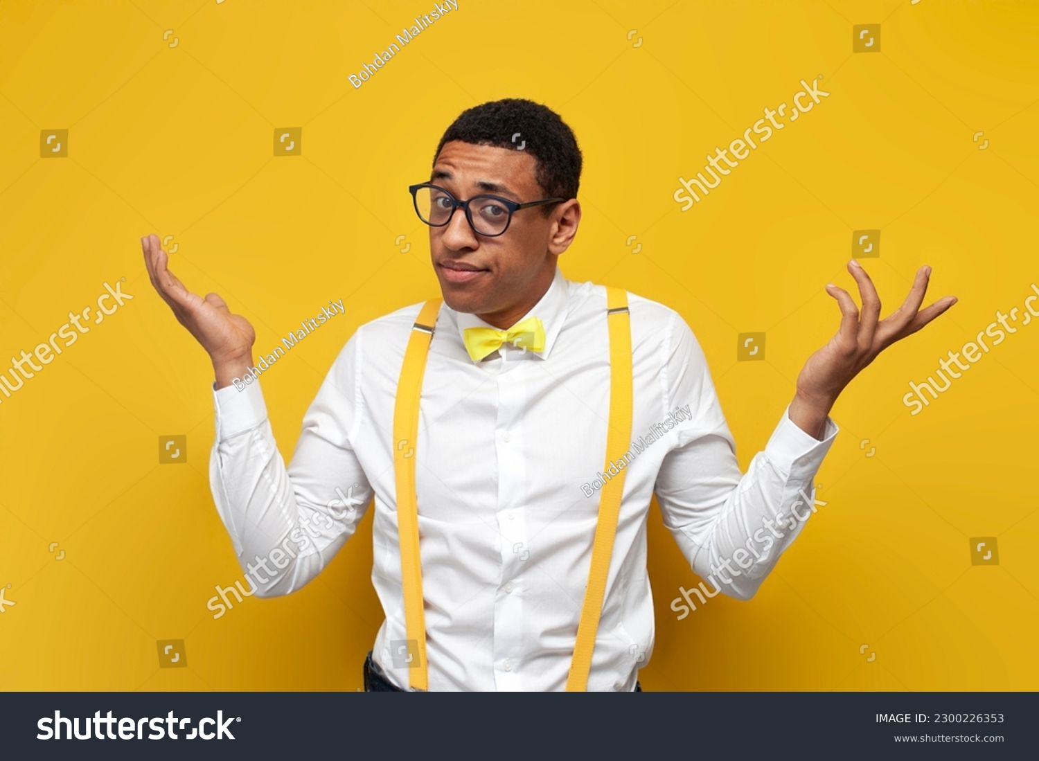 young insecure guy african american in white shirt with suspenders and bow tie spreads his arms on yellow isolated background, nerd man in glasses shrugs his shoulders #2300226353