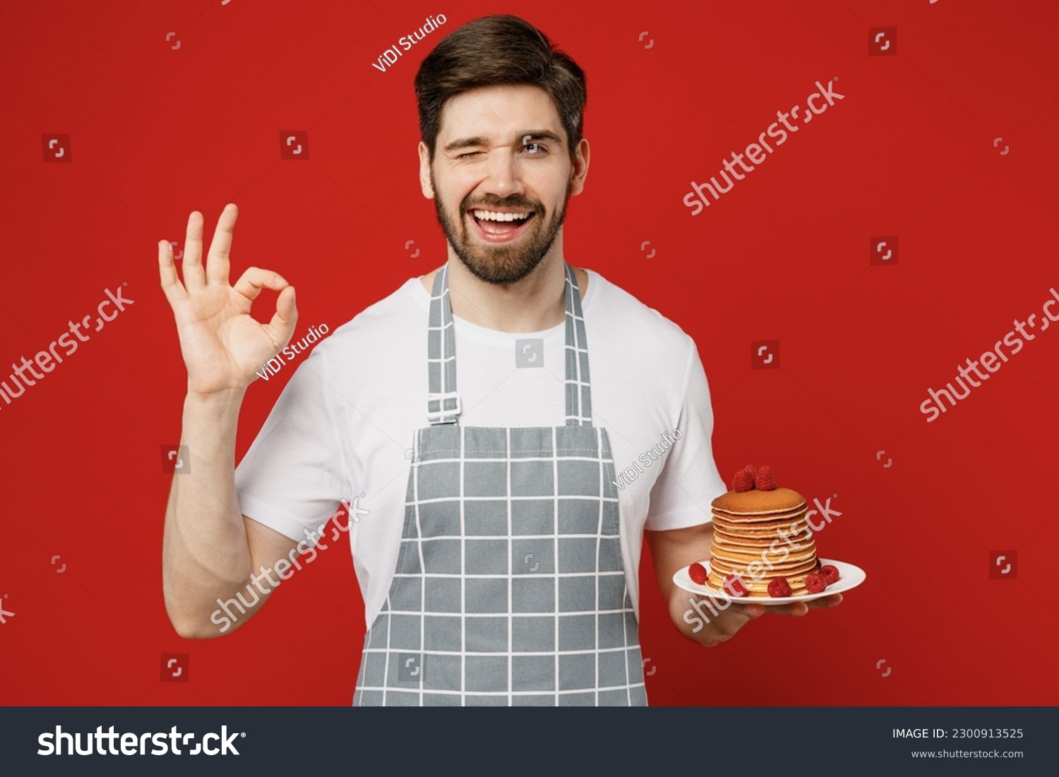 Young smiling happy fun male housewife housekeeper chef cook baker man wearing grey apron hold in hand plate with pancakes show ok gesture isolated on plain red background studio. Cooking food concept #2300913525